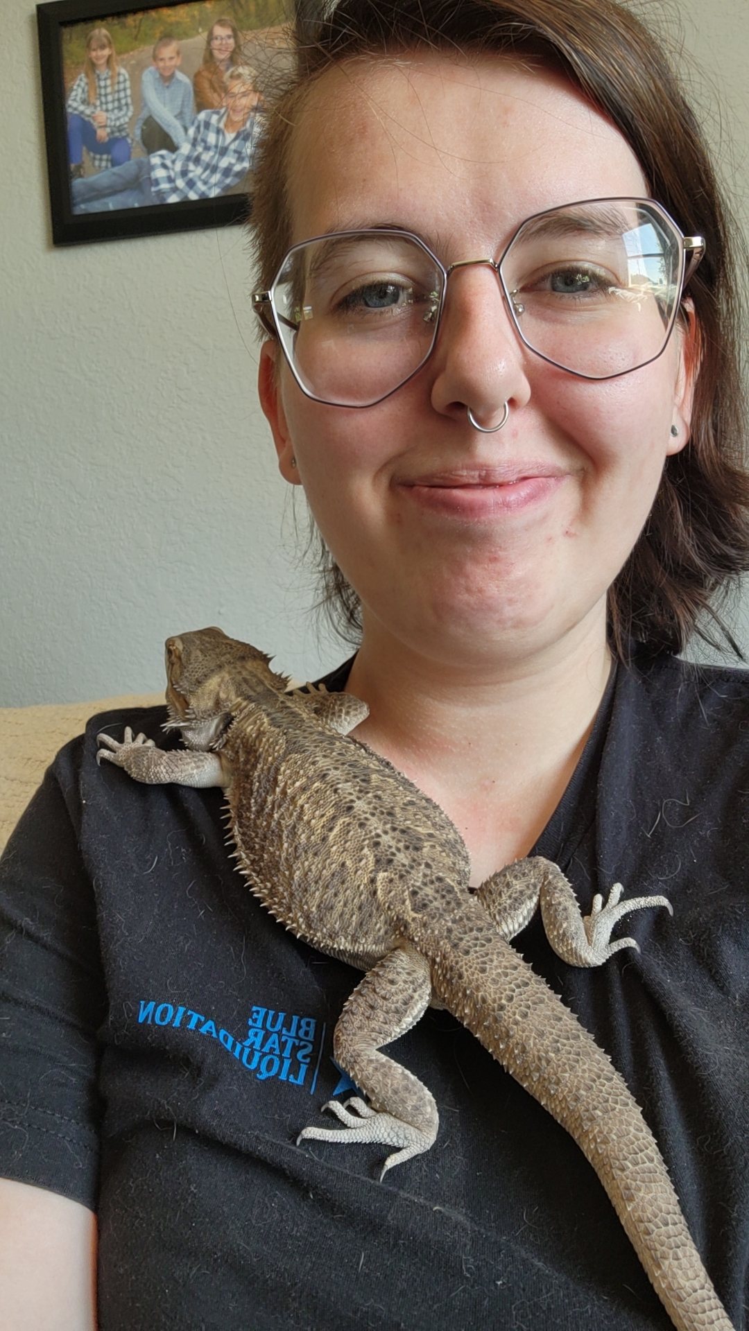 A photo of a girl with dark hair, with the left side shaved, large glasses, and a septum piercing with a bearded dragon on her chest.