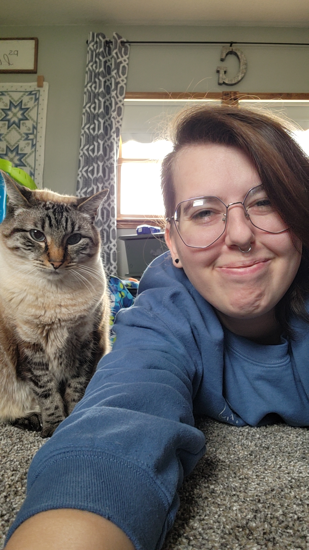 A photo of a girl with dark hair, with the left side shaved, large glasses, and a septum piercing laying on the ground next to a white and gray cat with blue eyes.