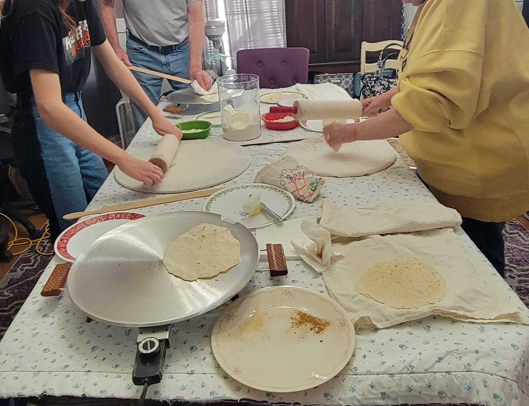 A picture of lefse tools. These tools include griddles, spatulas, plates, rolling pins, and rolling stones. There are three sets of arms in the photo as the people prepare lefse.