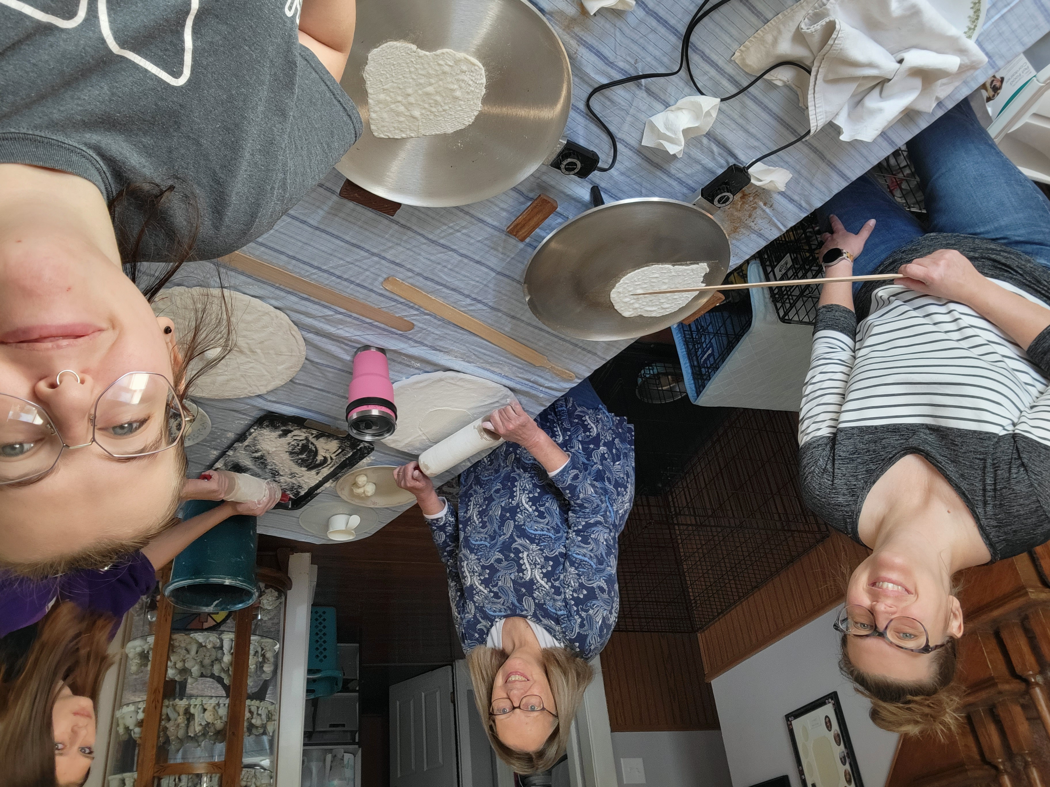 Myself, my mother, my sister, and my grandmother cooking lefse on the dining room table. On the table are the two lefse gridles, two rolling stones, two rolling pins, a cookie sheet of flour, and a few lefse paddles.